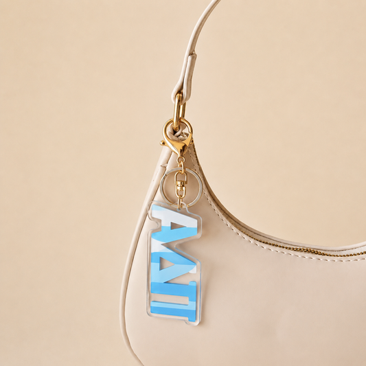 A cream-colored handbag with a gold zipper and The Darling Effect’s Alpha Delta Pi Stripes Acrylic Keychain—featuring a blue and white tag—makes a perfect, stylish sorority gift for statement-makers.