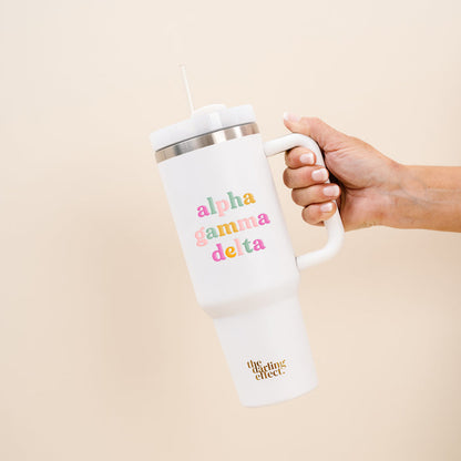 A hand holds The Darling Effect's Alpha Gamma Delta 40 oz Take Me Everywhere White Tumbler with a straw. The tumbler displays colorful "alpha gamma delta" text and gold "the uplifting effect" script. Background is solid light beige.