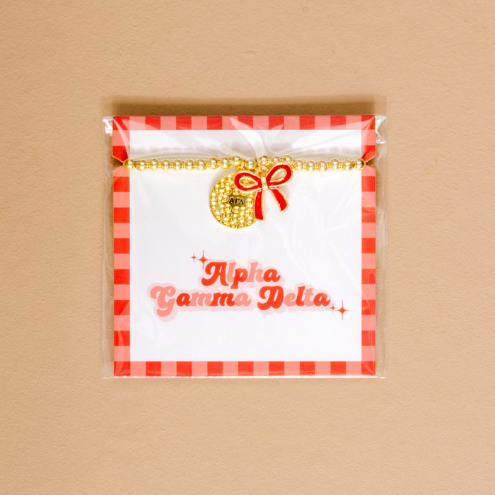 Show your Alpha Gamma Delta pride with The Darling Effect’s Bow Gold Charm Bracelet, featuring gold beads, a round charm, and red bow, displayed on a white card with a red checkered border and pink script.