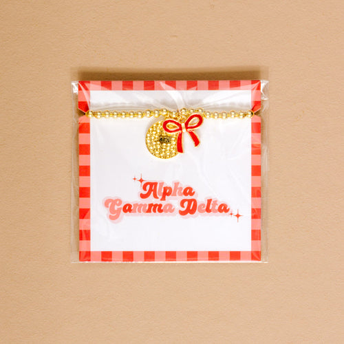 Show your Alpha Gamma Delta pride with The Darling Effect’s Bow Gold Charm Bracelet, featuring gold beads, a round charm, and red bow, displayed on a white card with a red checkered border and pink script.