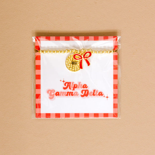 Show your Alpha Gamma Delta pride with The Darling Effect’s Bow Gold Charm Bracelet, featuring gold beads, a round charm, and red bow, displayed on a white card with a red checkered border and pink script.