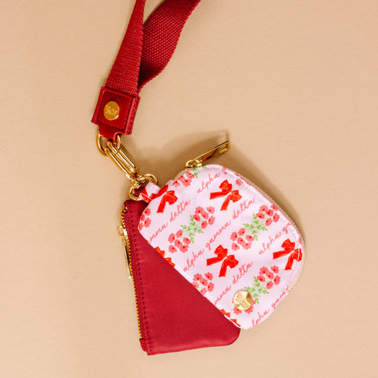 The Darling Effect Alpha Gamma Delta Dynamic Duo Pouch Wristlet features a zippered design with a red strap, gold clasp, and a red floral pattern with bows and "ahna yumna dekka" script on light pink.