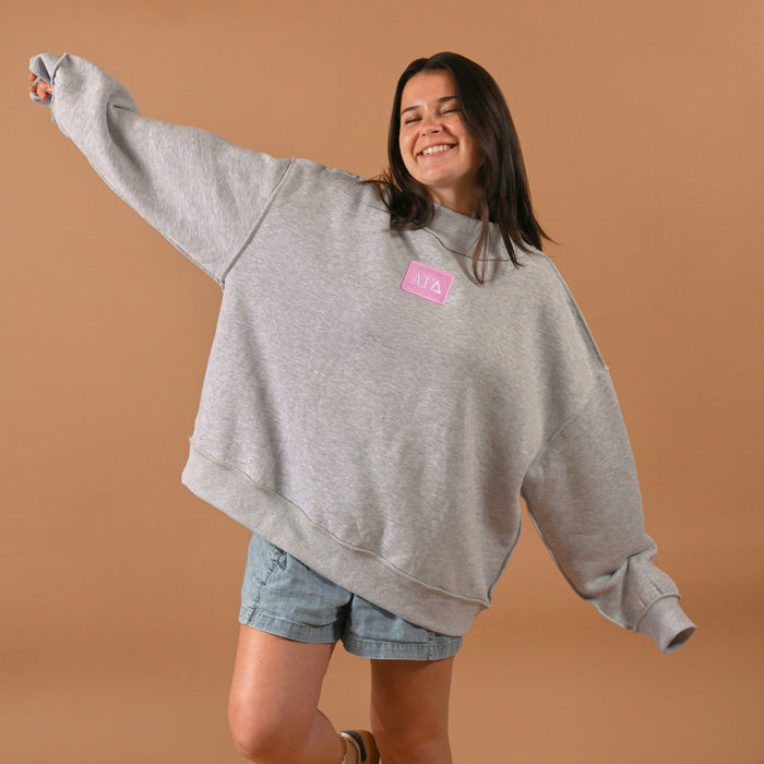 A smiling woman wearing The Darling Effect Alpha Gamma Delta Letter Patch Mockneck Sweatshirt (S/M) and denim shorts stands against a plain brown background with her arms outstretched in a joyful pose.