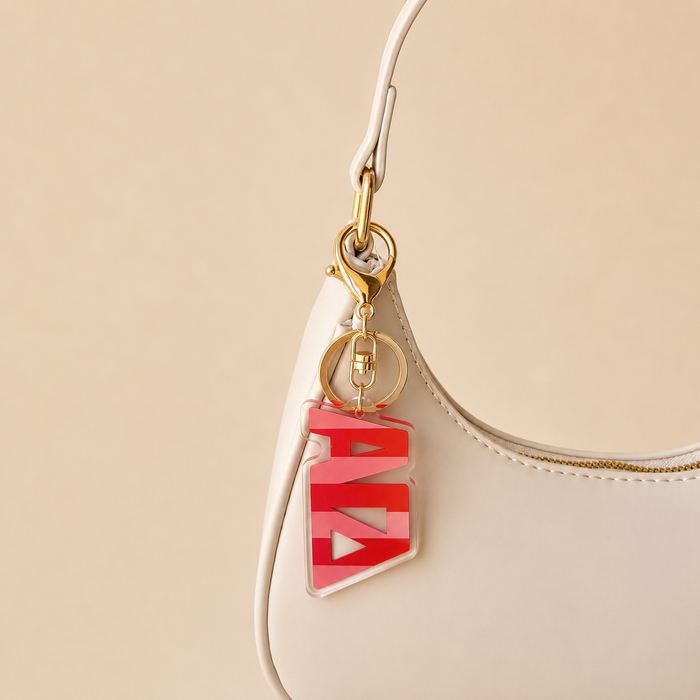 A cream-colored handbag with gold hardware features The Darling Effect's Alpha Gamma Delta Stripes Acrylic Keychain in a red and white geometric design on the handle. Set against a beige background, it makes a stylish sorority gift.