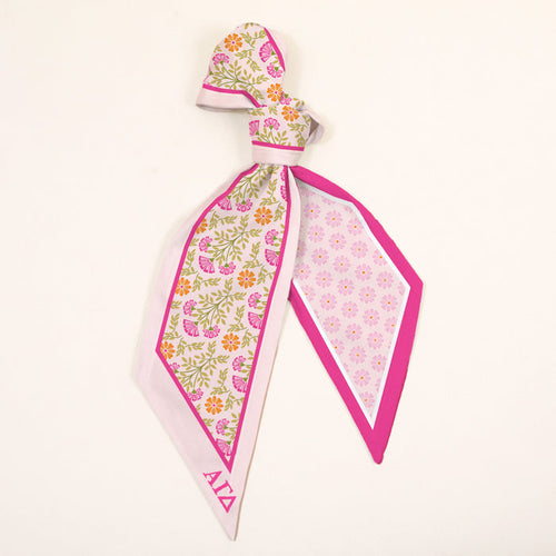 The Darling Effect’s Alpha Gamma Delta Twirl & Tie Scarf Accessory features pink, yellow, and green florals on one side, a pink geometric print on the other, pink borders, and elegant Alpha Gamma Delta letters in the corner.