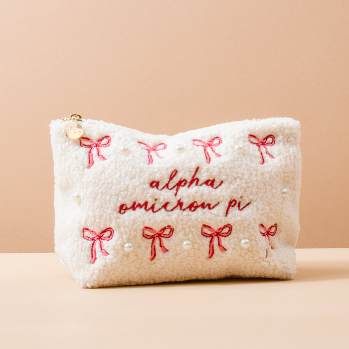The Darling Effect’s Alpha Omicron Pi Bows & Pearls Teddy Pouch features white sherpa, pink embroidered bows, pearl accents, and red “alpha omicron pi” stitching—a charming sorority gift shown on a beige background.