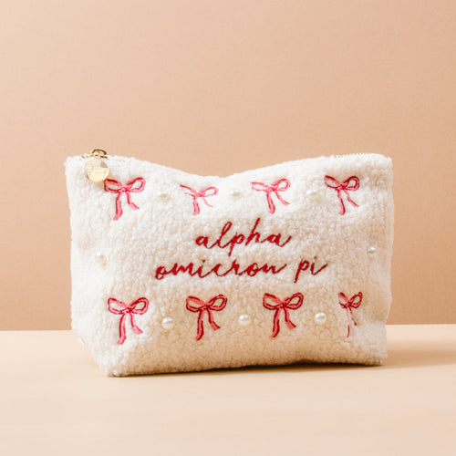 The Darling Effect’s Alpha Omicron Pi Bows & Pearls Teddy Pouch features white sherpa, pink embroidered bows, pearl accents, and red “alpha omicron pi” stitching—a charming sorority gift shown on a beige background.