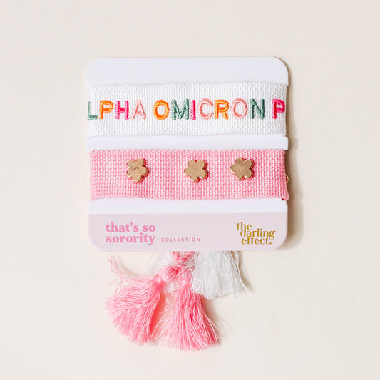 The Darling Effect’s Alpha Omicron Pi Woven Bracelet Set features three bracelets—white with “ALPHA OMICRON PI” embroidery, pink with gold flower charms, and pink with white tassels—ideal for bid day or as a sisterhood gift.