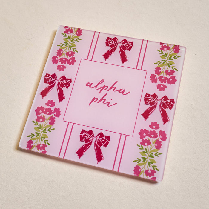 The Darling Effect’s Alpha Phi Acrylic Coaster features a pink striped background with “alpha phi” in cursive, accented by red bows and pink flowers with green leaves—an ideal sorority gift.
