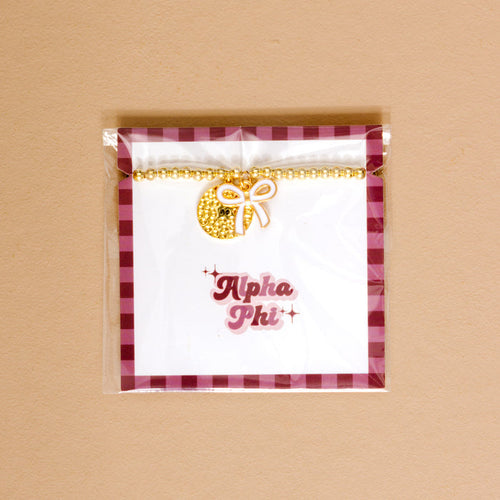 The Darling Effect’s Alpha Phi Bow Gold Charm Bracelet features a gold beaded design with a round textured charm and bow, packaged on an Alpha Phi card with pink letters and a checkered border—perfect for showing chapter pride.