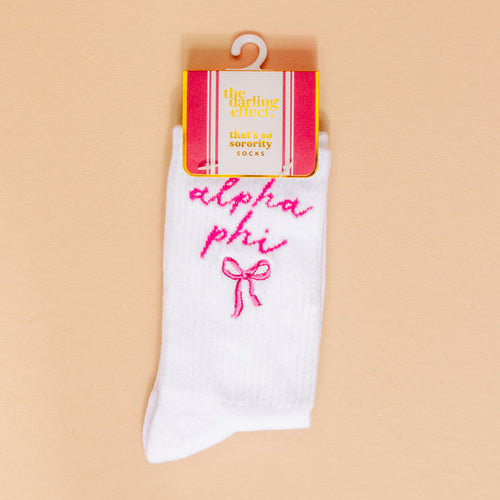 Alpha Phi Embroidered Crew Socks by The Darling Effect feature pink "Alpha Phi" embroidery and a pink bow. Packaged in cute pink and white with "the darling effect" text, they're ideal sorority gifts. Light peach background.