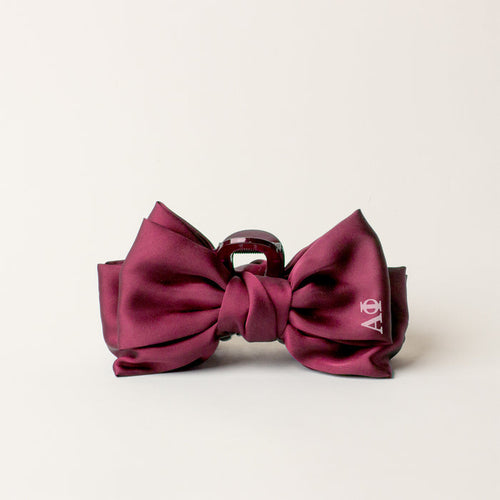 The Darling Effect Alpha Phi Satin Bow Claw Clip in maroon features glossy satin fabric and a small white logo on one bow, making it an elegant hair accessory for updos. Displayed against an off-white background.