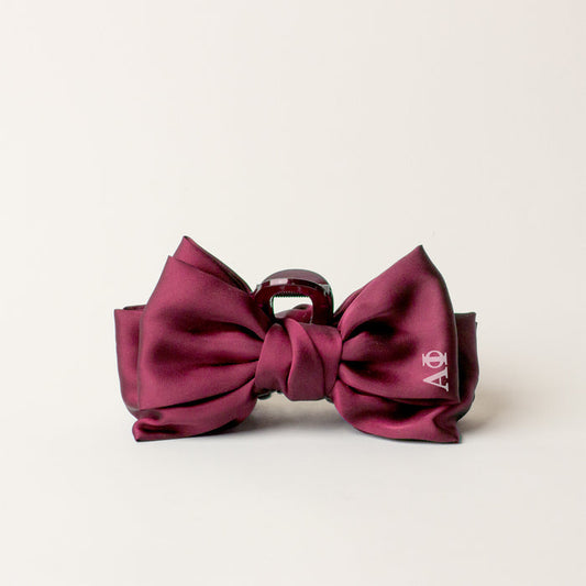 The Darling Effect Alpha Phi Satin Bow Claw Clip in maroon features glossy satin fabric and a small white logo on one bow, making it an elegant hair accessory for updos. Displayed against an off-white background.