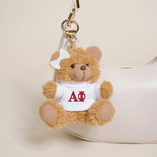 The Darling Effect Alpha Phi Teddy Bear Bag Charm features a small plush bear with a white bow and Greek letters ΑΦ on its shirt, hanging as a cute keychain from a handbag.