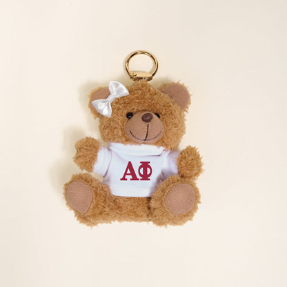 The Darling Effect’s Alpha Phi Teddy Bear Bag Charm is a small plush bear with a white bow and shirt featuring red ΑΦ letters, perfect as a cute sorority gift or accessory for your bag.