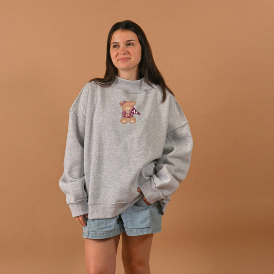 A woman poses against a tan background, wearing The Darling Effect's Alpha Phi Teddy Bear Mockneck Sweatshirt (L/XL) in gray with a bear graphic, paired with light denim shorts and a subtle smile, her hands tucked in her pockets.