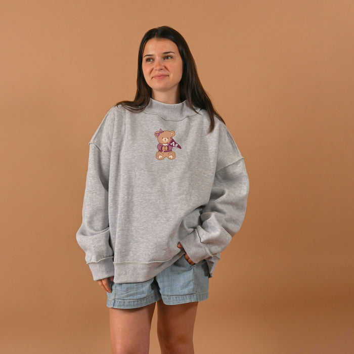A woman wearing The Darling Effect's Alpha Phi Teddy Bear Mockneck Sweatshirt S/M and light denim shorts stands against a beige background, smiling slightly with her hands in her pockets.