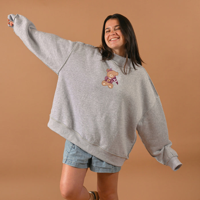 A young woman with eyes closed smiles, wearing The Darling Effect's Alpha Phi Teddy Bear Mockneck Sweatshirt (S/M) and blue denim shorts, standing against a brown background with one arm outstretched.