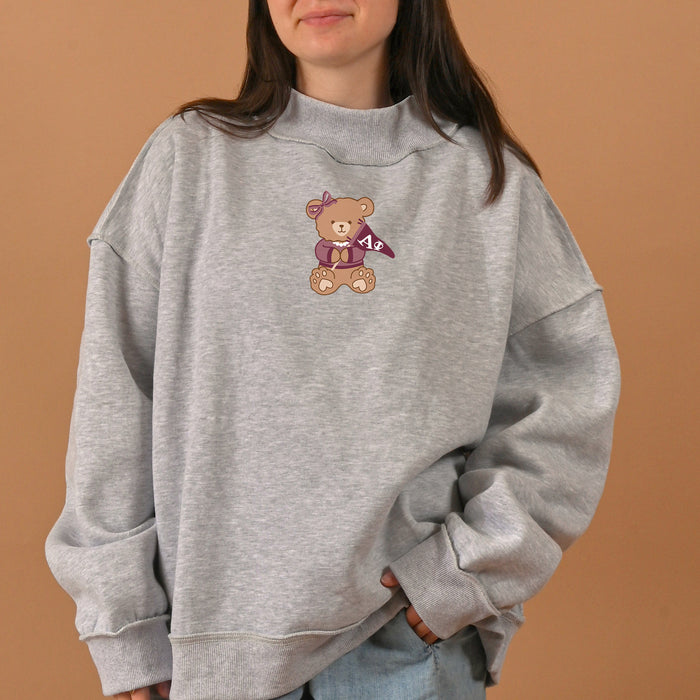 A woman wearing The Darling Effect's Alpha Phi Teddy Bear Mockneck Sweatshirt in size S/M, an oversized gray sweatshirt featuring a teddy bear graphic holding a purple pennant with "AΦE" on the front. The background is a plain tan color.