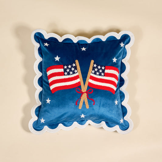 The Darling Effect’s American Flags Scalloped Square Throw Pillow showcases two crossed flags and white stars embroidered on a neutral background with blue fabric and scalloped white edges.