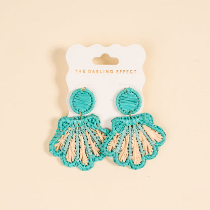The Darling Effect's Aqua Cabana Breeze Earrings—turquoise and beige woven shells on a white card with the brand print—are lightweight, boho-chic accessories perfect for everyday style against a light beige backdrop.