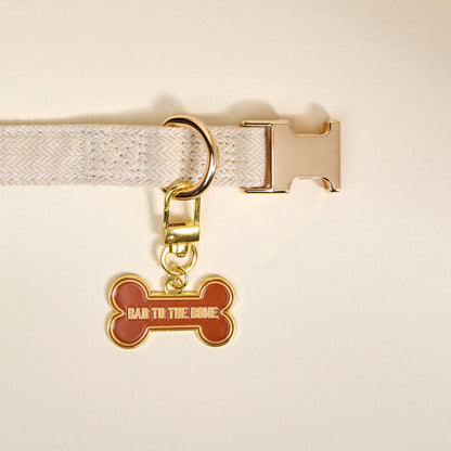 The Darling Effect’s Bad to the Bone Pawfect Collar Charm, a beige collar with gold buckle and a bone-shaped tag reading "BAD TO THE BONE" in gold letters, adds playful flair to your pet accessories collection. Displayed on a light background.