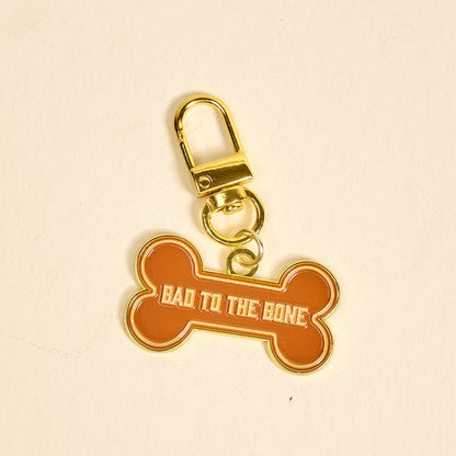 The Darling Effect’s Bad to the Bone Pawfect Collar Charm features an orange dog bone shape with gold edges, a gold clasp, and “BAD TO THE BONE” in bold white letters—a fun accessory for your pet’s collar or keychain.
