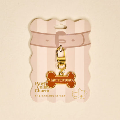 The Darling Effect’s Bad to the Bone Pawfect Collar Charm features a brown bone shape with gold trim and "BAD TO THE BONE" text, attached to gold hardware and displayed on a pink and white card with collar, cat, and dog icons.