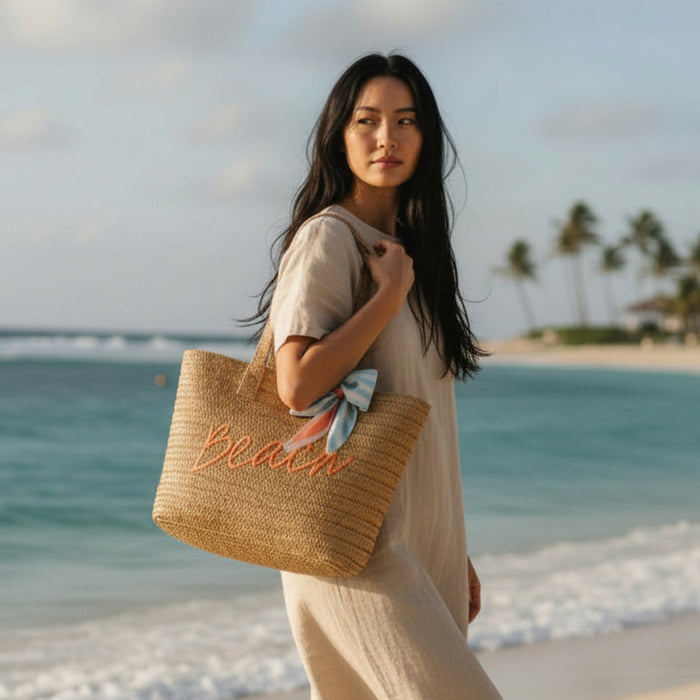 A woman in a beige dress stands on a sandy beach holding The Darling Effect's Beach Straw & Sun Tote, featuring "Beach" lettering and a blue scarf tied to the handle. Palm trees sway nearby, creating the perfect vacation vibe.