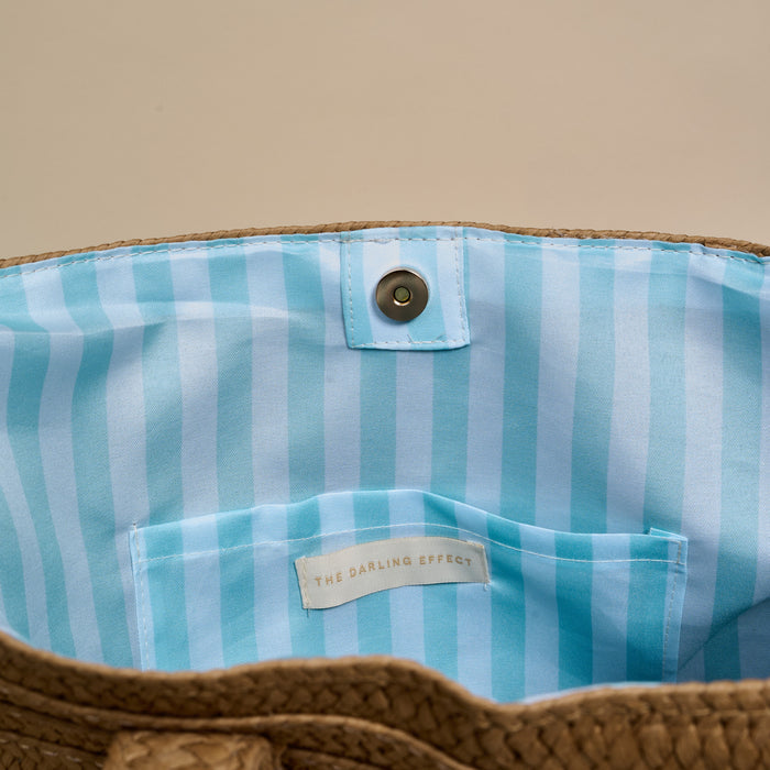 The image displays the inside of The Darling Effect's Beach Straw & Sun Tote, featuring a blue and white striped lining, snap closure, and a small branded interior pocket.