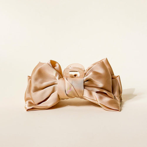 The Darling Effect Beige Satin Bow Claw Clip features a large, shiny satin bow on a clear plastic clip, shown on a cream background—perfect for adding elegance to any updo.