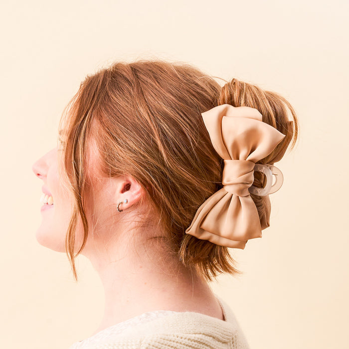 A woman with light brown hair in an updo, secured by The Darling Effect's Beige Satin Bow Claw Clip, smiles while facing left against a light cream background. She wears a cream top and small hoop earrings.