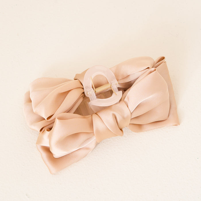 The Darling Effect Beige Satin Bow Claw Clip features a beige satin bow on a translucent light pink base, ideal for enhancing any updo, and is pictured on a cream-colored surface.