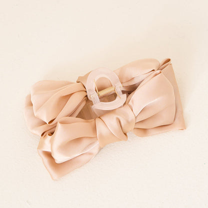 The Darling Effect Beige Satin Bow Claw Clip features a beige satin bow on a translucent light pink base, ideal for enhancing any updo, and is pictured on a cream-colored surface.
