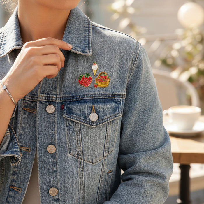 A person in a light blue denim jacket adorned with The Darling Effect’s Berrylicious The Pin Pack—featuring a strawberry, ice cream cone, and picnic basket—sits at an outdoor table with a blurred cup and saucer in the background.