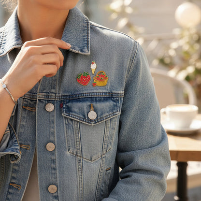 A person in a light blue denim jacket adorned with The Darling Effect’s Berrylicious The Pin Pack—featuring a strawberry, ice cream cone, and picnic basket—sits at an outdoor table with a blurred cup and saucer in the background.
