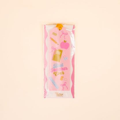 The Darling Effect's Best Teacher Ever Acrylic Bookmark features pink and white colors, school-themed illustrations, and “Best Teacher Ever” text. It comes in clear plastic packaging with a yellow ribbon, making it a perfect gift for book lovers.