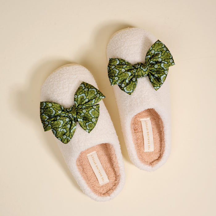 The Darling Effect’s Betty Bow Everbloom Forest Green Fuzzy Slippers feature a cozy cream exterior, soft pink inner lining, and green bows with a white floral pattern, shown on a light beige background.