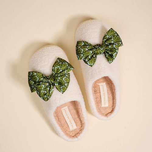 The Darling Effect’s Betty Bow Everbloom Forest Green Fuzzy Slippers feature a cozy cream exterior, soft pink inner lining, and green bows with a white floral pattern, shown on a light beige background.