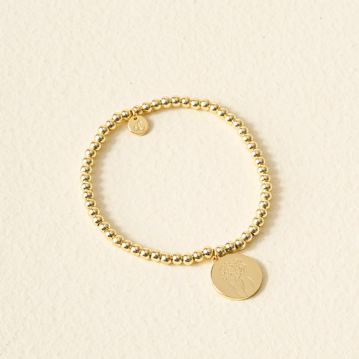 The Darling Effect's Birth Flower Beaded Charm Bracelet features two round gold charms—one engraved with a flower—on a gold beaded band, displayed on a textured surface; an ideal personalized jewelry gift or birth flower bracelet.