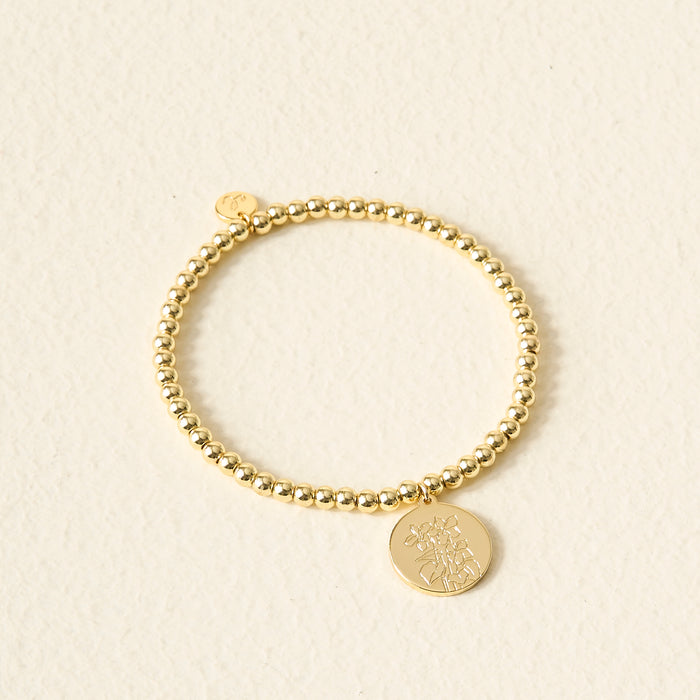 The Darling Effect Birth Flower Beaded Charm Bracelet features a gold beaded design with a round engraved floral charm, displayed on a cream textured surface—a stylish piece of personalized jewelry for any occasion.