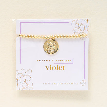 The Darling Effect Birth Flower Beaded Charm Bracelet features a gold beaded design with a round floral charm, displayed on a "Month of February, violet" card accented in gold and purple—perfect for personalized jewelry lovers.