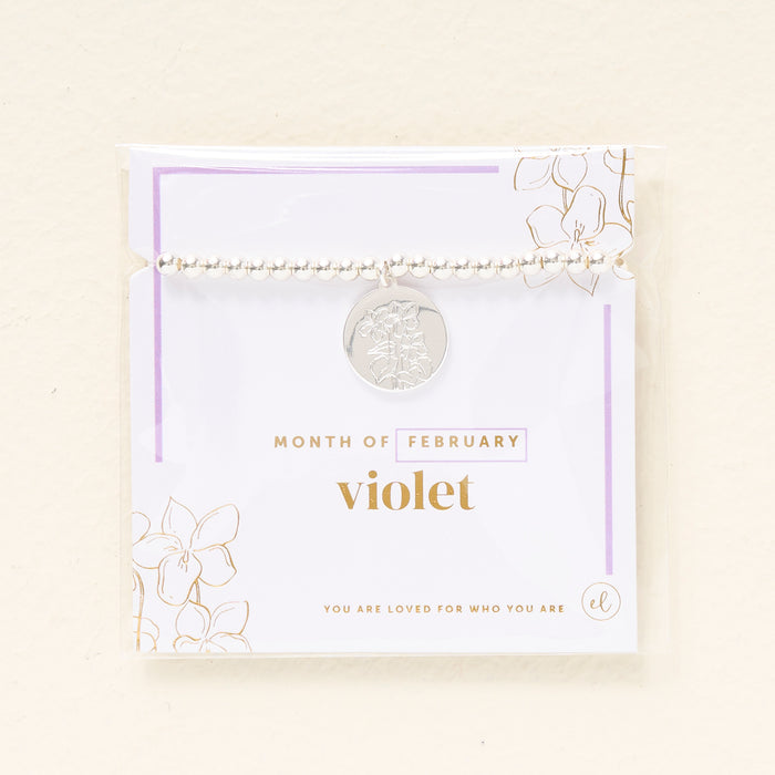 The Darling Effect Birth Flower Beaded Charm Bracelet features a silver beaded bracelet with a violet charm on a February card, adorned with gold and lavender florals and the message "You are loved for who you are.