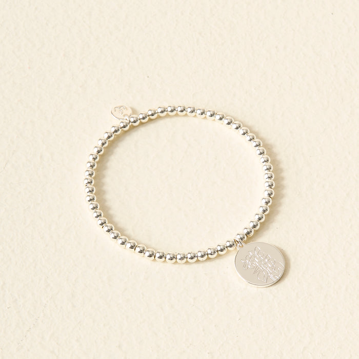 The Darling Effect Birth Flower Beaded Charm Bracelet, featuring a silver beaded band and round engraved charm, rests on a light textured surface—an ideal personalized jewelry piece for your style.