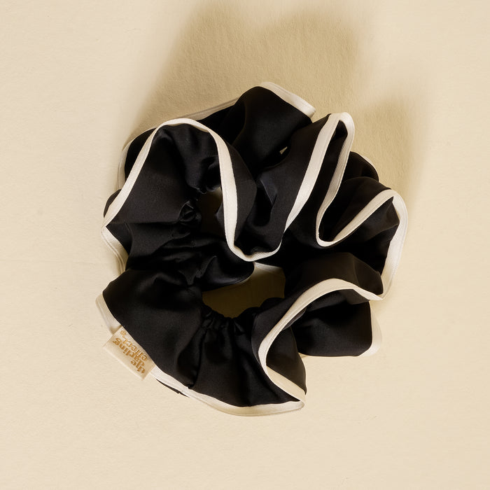 The Darling Effect's Black Ruffle Up Scrunchie, featuring white trim and a small beige tag, is arranged in a circle on a beige background.