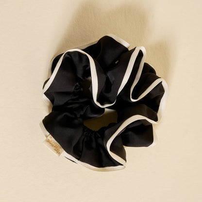 The Darling Effect's Black Ruffle Up Scrunchie, featuring white trim and a small beige tag, is arranged in a circle on a beige background.