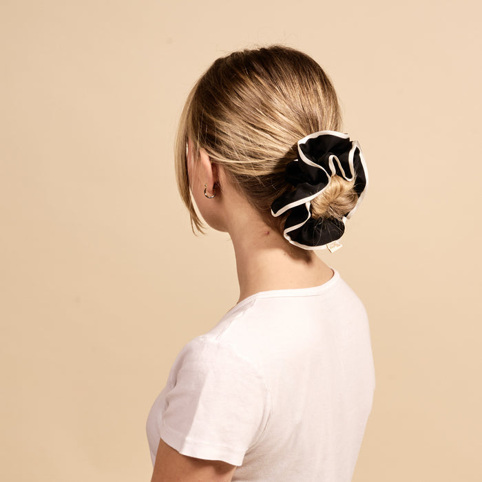 A woman with blonde hair in a low bun models The Darling Effect’s Black Ruffle Up Scrunchie, an oversized black accessory with white trim. She wears a plain white t-shirt and faces away from the camera against a beige background.