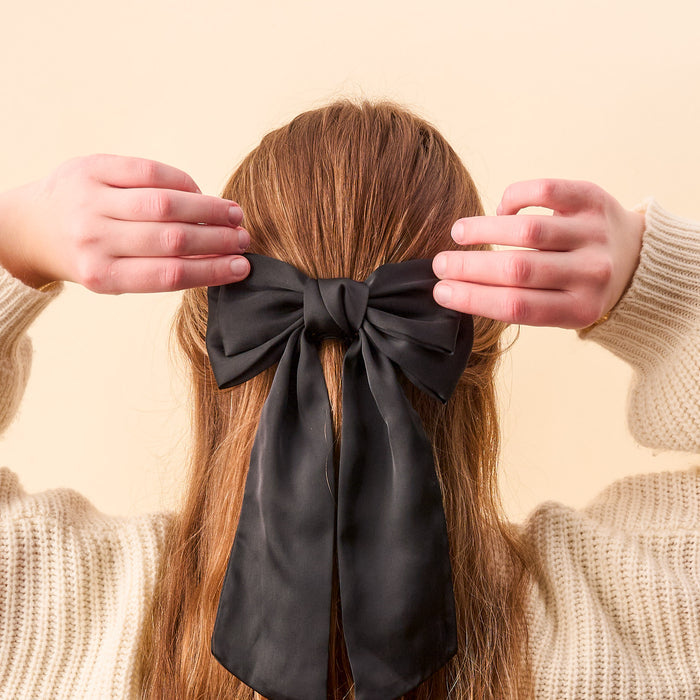 A person with long brown hair, seen from behind, wears The Darling Effect’s Black Satin Hair Bow. They are dressed in a cream-colored sweater against a light beige background, highlighting this elegant hair accessory.