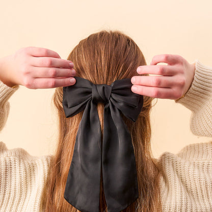 A person with long brown hair, seen from behind, wears The Darling Effect’s Black Satin Hair Bow. They are dressed in a cream-colored sweater against a light beige background, highlighting this elegant hair accessory.
