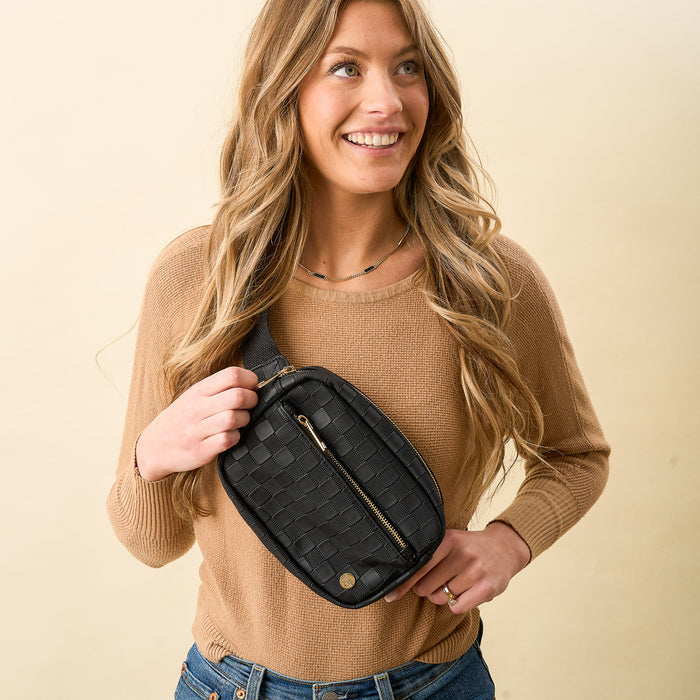 A smiling woman with long wavy blonde hair wears a tan sweater and jeans, holding The Darling Effect’s Black Urban Check Belt Bag across her chest, standing against a plain beige background.
